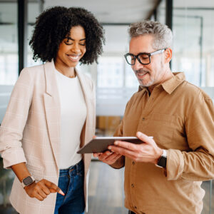 Two colleagues smilingly look at a tablet in a bright office. The woman wears a plaid blazer, and the man, with glasses, wears a beige shirt. Collaborative atmosphere.