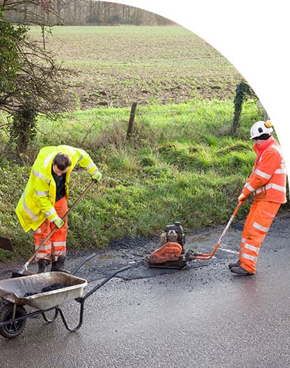 Public works crew in high-visibility gear repairing a roadway, with one worker pouring material from a wheelbarrow while another operates paving equipment along a rural roadside.