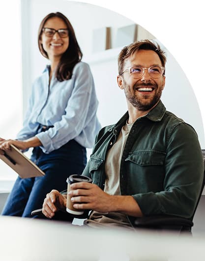Two people in casual attire smile in a bright office. A man in glasses holds a coffee, while a woman with a tablet sits on a desk, conveying positivity.
