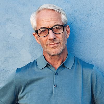 Adult professional white male with combed white hair wearing a blue polo shirt and black glasses while standing against a blue wall
