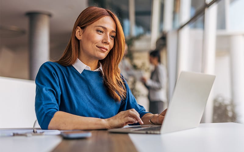 A focused red-haired woman in a blue sweater works on a laptop at a table in a bright, modern office, conveying calm confidence and productivity.