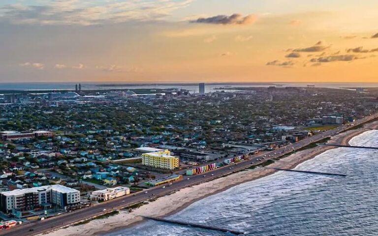 Aerial view of Galveston, TX at sunset, showing ocean waves along a sandy beach, a beachfront road with colorful buildings, and a dense neighborhood stretching toward the horizon.