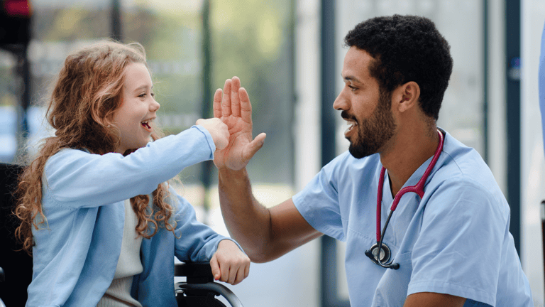 A young girl in a wheelchair enthusiastically gives a high five to a friendly male doctor in a well-lit medical setting.
