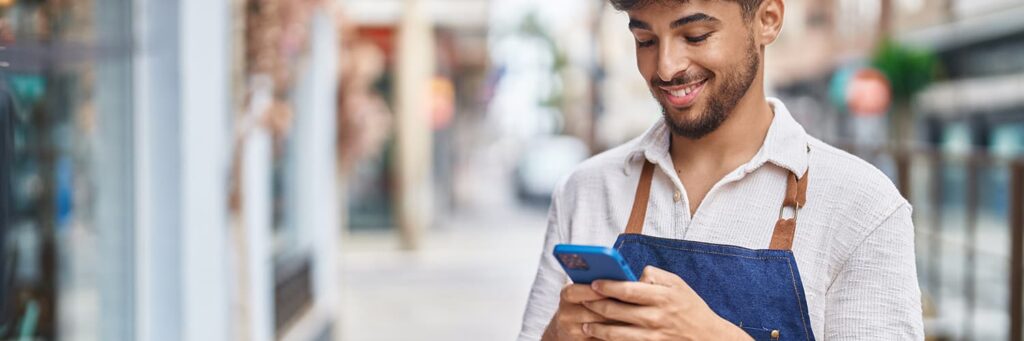 Young mixed race male business owner smiling while using his blue phone outside of a small business