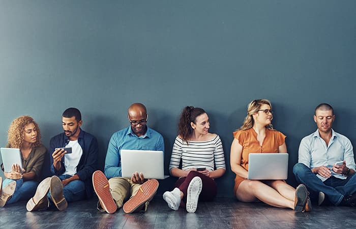 A diverse group of public sector employees sitting on the floor and sharing engagement tips & using community engagement tools on different devices.