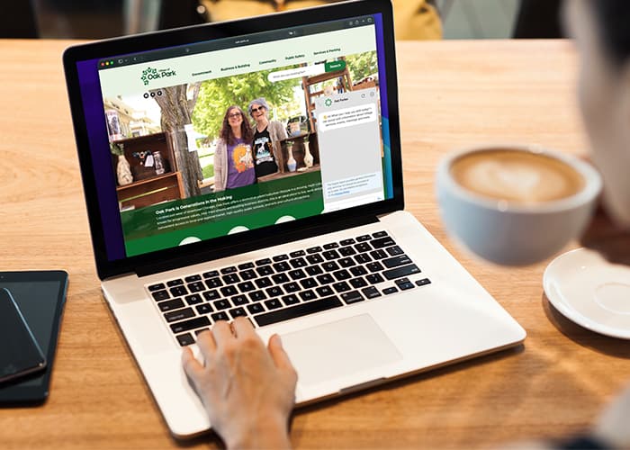 MacBook Pro displaying the Oak Park government website with the Oak Parker AI chat assistant open while a person drinks coffee in the background.