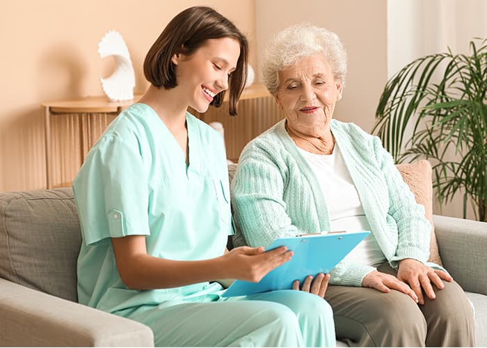 Young nurse with clipboard and senior woman sitting on sofa at home