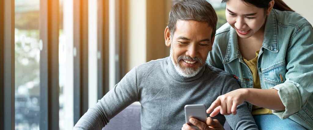 Asian male using his cell phone for a personalized enrollment experience while his adult daughter smiles and points to a displayed benefit, symbolizing improved health and human services accessibility.