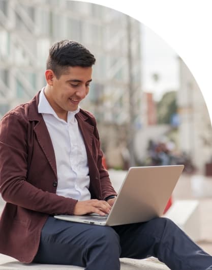 Person wearing a maroon blazer and white shirt, sitting outdoors and typing on a silver laptop with modern buildings in the background.