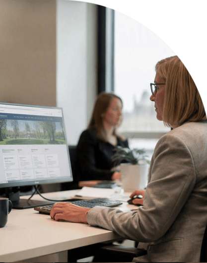 Person seated at a desk using a computer in an office, with a colleague blurred in the background near a window.