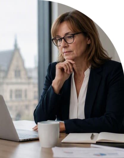Person wearing glasses working on a laptop at a desk with a notebook and coffee cup beside a window.