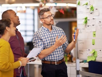 A man standing in front of a whiteboard full of post-it notes, explaining a scenario to his colleagues.