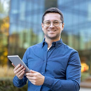 Smiling man in glasses and a blue shirt holds a tablet outside a modern glass office building.