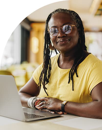 Mature woman with dreadlocks and glasses, wearing a yellow top, smiles while sitting at a laptop in a bright, cozy cafe. The tone is warm and welcoming.