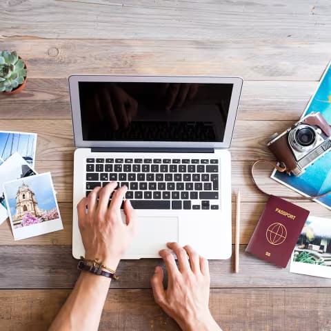 Hands typing on a laptop on a wooden table with travel photos, passport, vintage camera, and a succulent, conveying a travel planning theme.