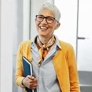 An older adult with short white hair and glasses smiles warmly, wearing a yellow cardigan and scarf. They're holding a book in a bright, modern government office.