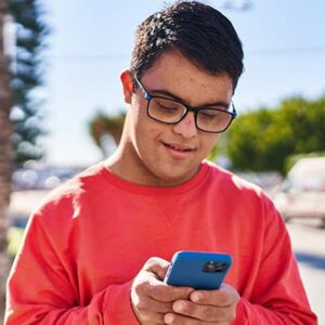 Young man in glasses and a red sweatshirt smiling while submitting a request for government assistance on his smartphone. He is outdoors on a sunny day, with trees and a blurred background.
