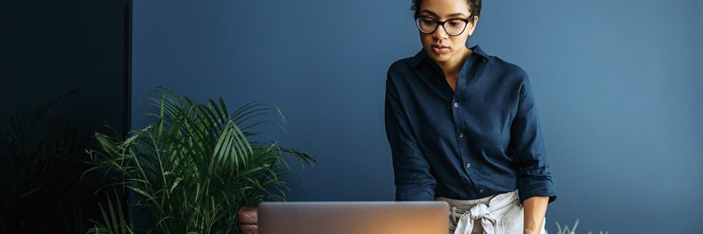 Young professional woman watching engaging content on her laptop, standing in an office with plants and a deep blue wall