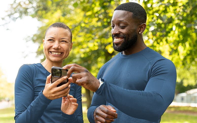 African American man and woman in a park smiling while looking at their mobile device