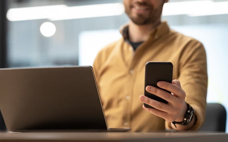Person using a smartphone while working on a laptop at a desk.