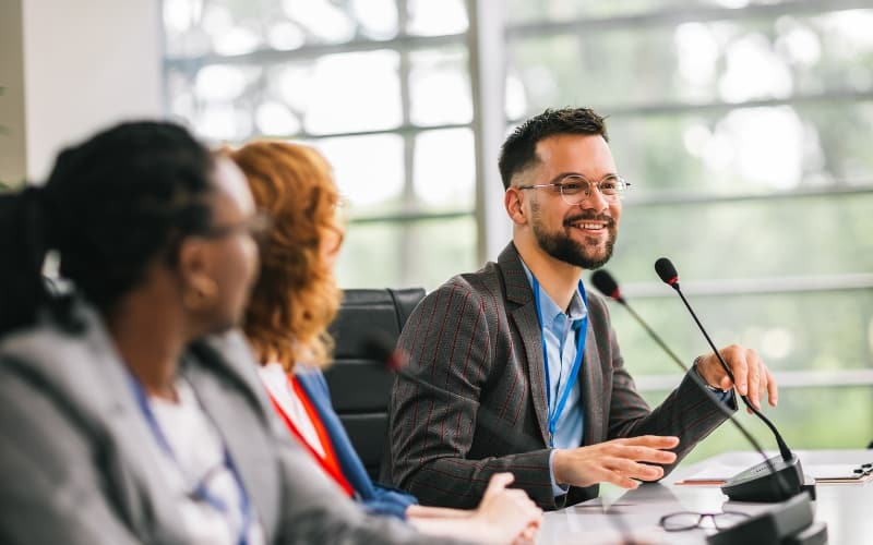 A council member speaks to residents during a meeting, seated at a conference table with microphones in a naturally lit room.