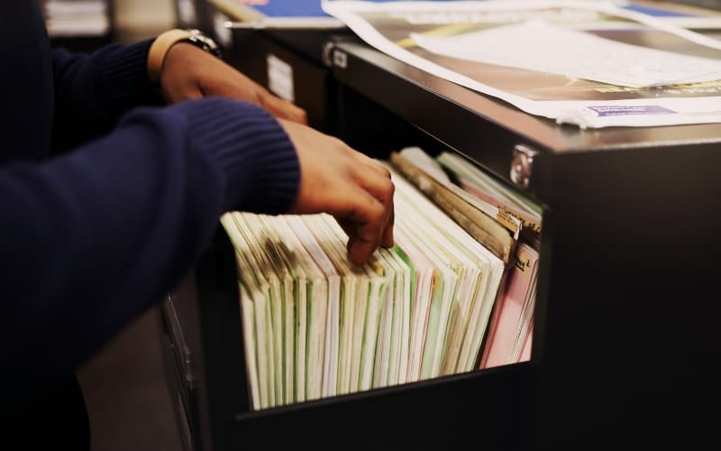 A person organizes folders in a black filing cabinet drawer, with papers and additional folders visible on top of the cabinet representing a search for public records.