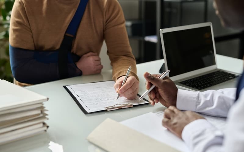 Healthcare professional assists an injured patient with Medicaid-related paperwork at a desk filled with documents and a laptop.