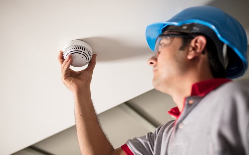 Worker in safety gear inspecting a smoke detector, emphasizing fire safety in short-term rentals.