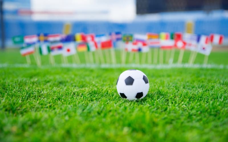 Soccer ball on green grass with multiple small international flags in the background, representing World Cup 2026.