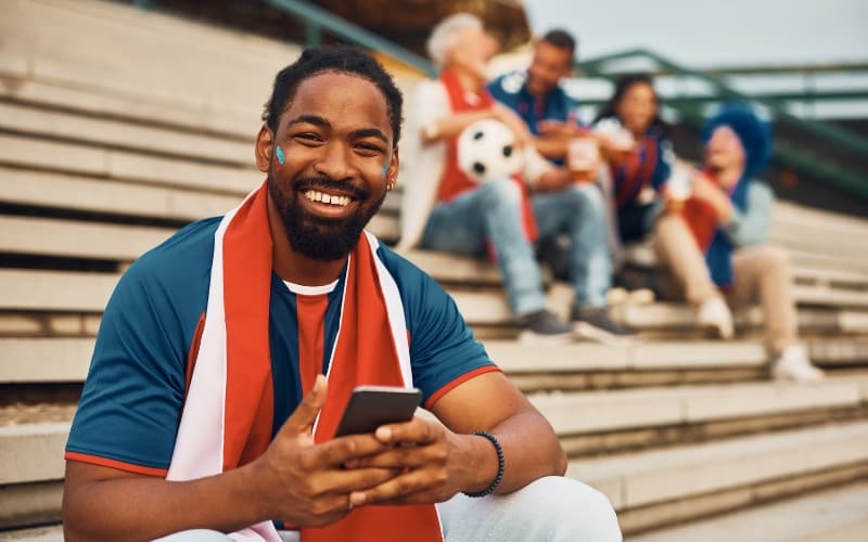 Soccer fan using a smartphone in stadium stands, highlighting the role of digital and multilingual engagement during World Cup 2026.