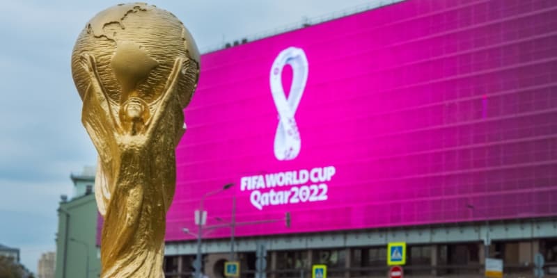 Golden FIFA World Cup trophy in front of a large pink billboard displaying the Qatar 2022 logo.