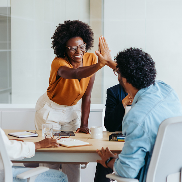 A woman in a brown top enthusiastically high-fives a seated man during a meeting. The scene conveys teamwork and positivity in a modern office setting.