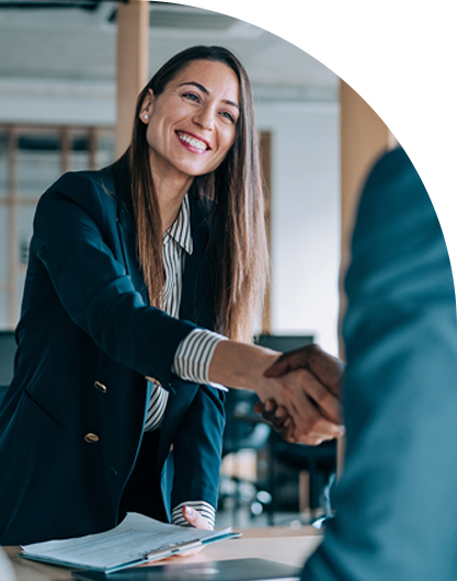 A woman in a blue blazer smiles warmly while shaking hands across a table in an office, conveying professionalism and friendliness.