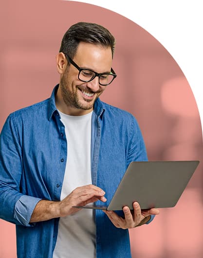 Smiling public sector communications professional with glasses using a laptop, wearing a blue shirt over a white tee. The blurred pink background adds warmth and a professional tone.