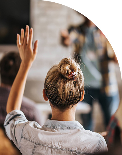 A woman with blonde hair in a bun raises her hand in a classroom setting. She wears a light denim shirt, while a blurred figure stands in the background. The tone is engaged and participatory.