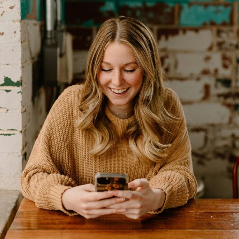 Smiling woman with long blonde hair in tan sweater, uses smartphone at wooden table in rustic cafe setting. Bright, cozy atmosphere.