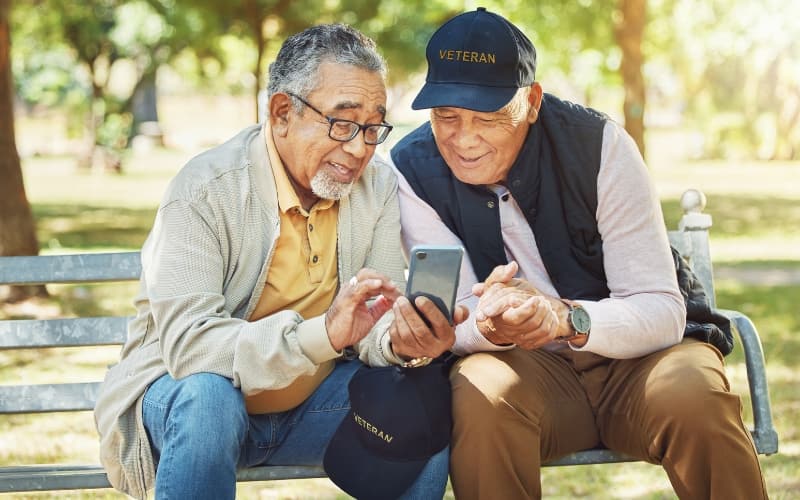 Two veterans wearing caps sit on a park bench, smiling and looking at a smartphone together in a sunny, green outdoor setting.
