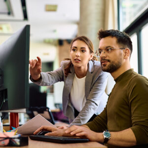 A woman in a grey blazer points at a computer screen, discussing with a man typing at a desk. They appear focused and engaged in a modern office.