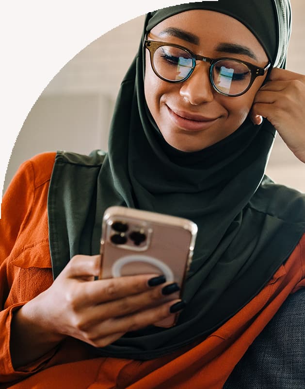 Young beautiful smiling woman in hijab while reading an SMS text message on her phone phone and sitting on the sofa