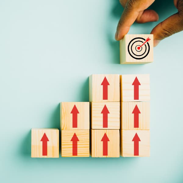 Wooden blocks form ascending steps with red upward arrows; a hand places a target-marked block on top, symbolizing growth and progress.